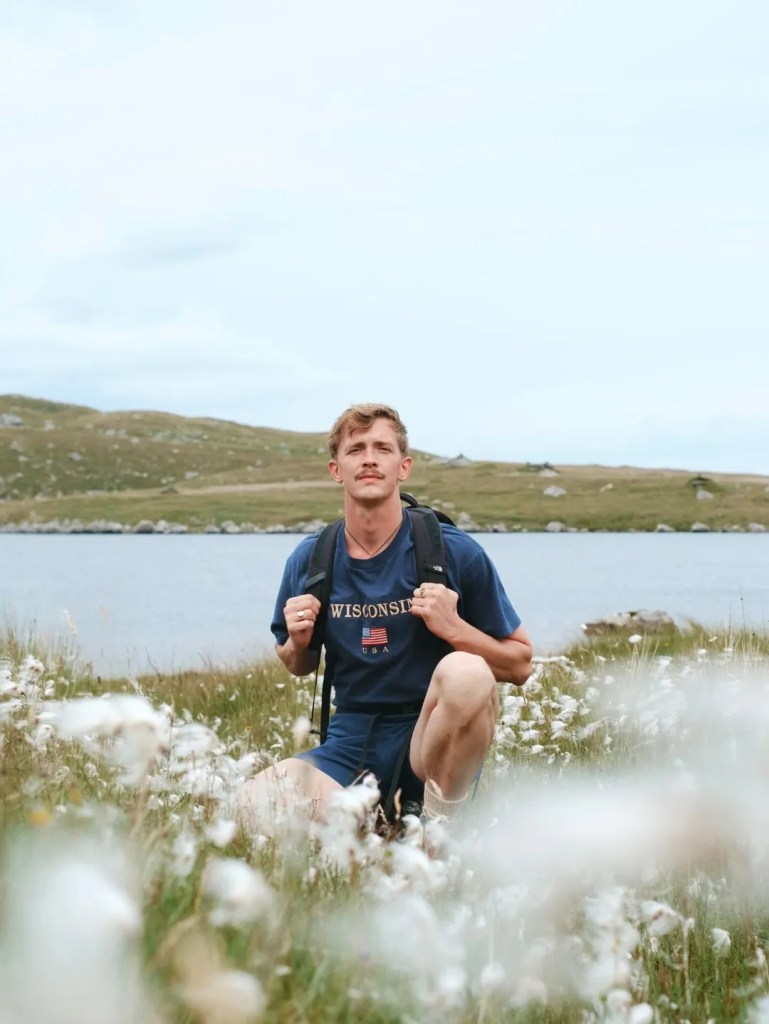 Stephen agenouillé au milieu d'un champ de fleurs blanches, un lac derrière lui. Porter un t-shirt et un short bleu marine foncé