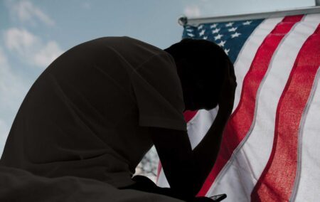 A distressed man in silhouette sits in dark room texting on smartphone. Blinds drawn in window light making silhouette of man with hand of forehead communicating on cell phone. Moody dark image with copy space.