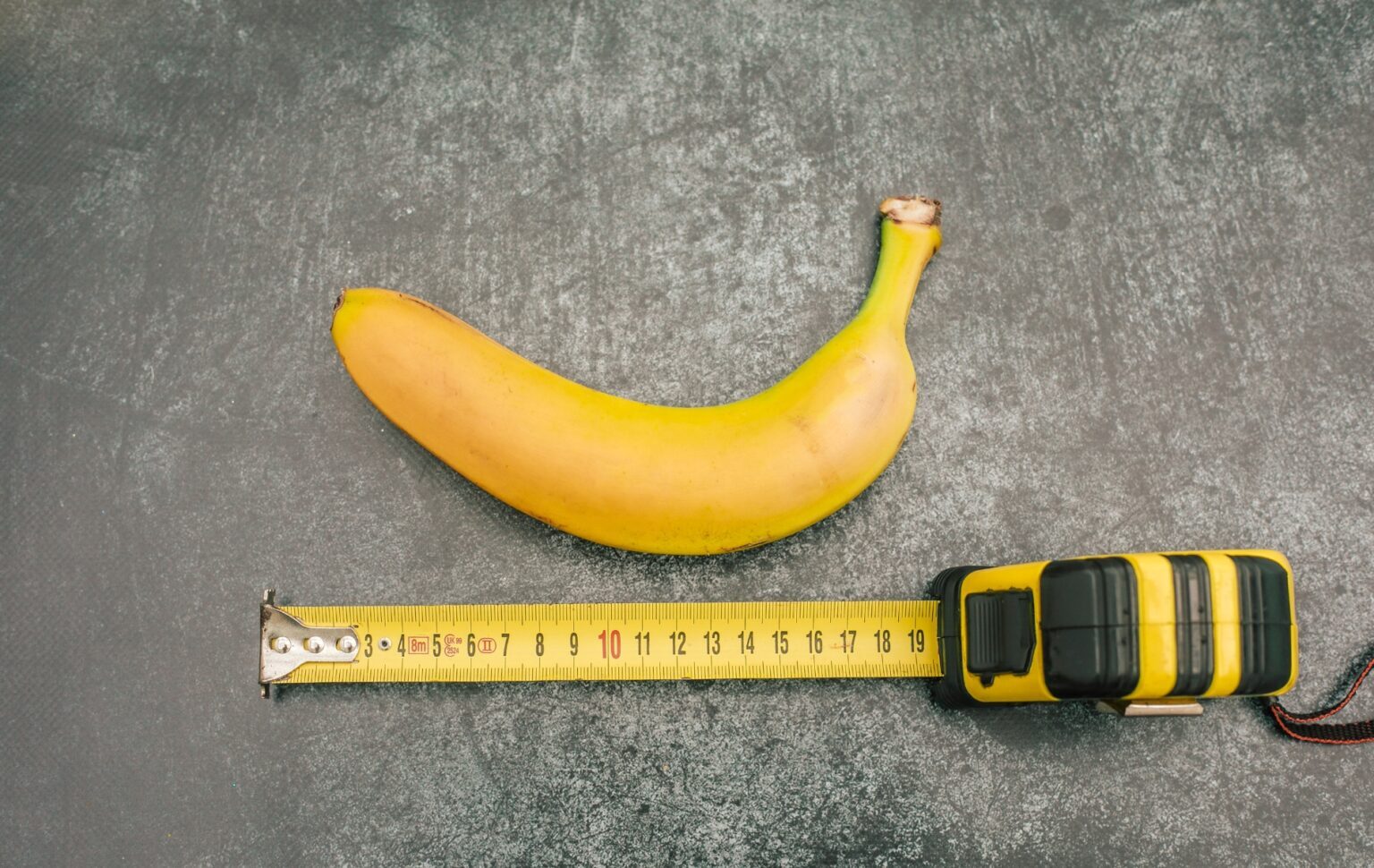 Ce sont les États américains avec la taille moyenne du pénis la plus grande – et la plus petite. banana on a grey table with a measuring tape below it