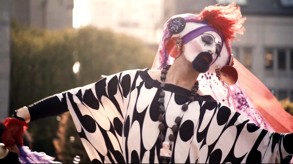 Hunky Jesus still : une personne habillée en drag avec une robe longue noire et blanche et un casque violet et orange.