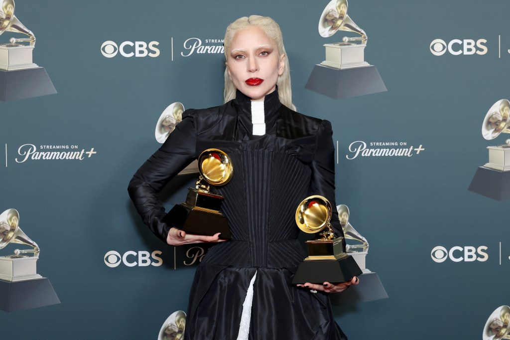 Lady Gaga debout avec deux Grammys dans une longue robe noire avec des détails blancs en dessous.