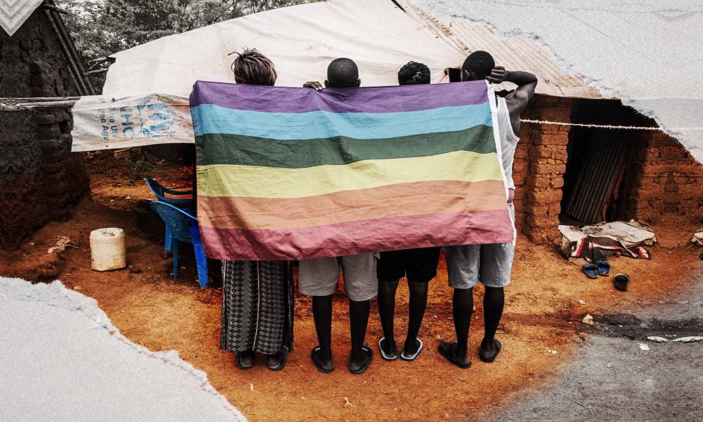 Une image montre quatre demandeurs d’asile debout, dos tournés à la caméra, dans le camp de réfugiés de Kakuma, au Kenya. Ils tiennent un drapeau de la fierté drapé sur leur dos.