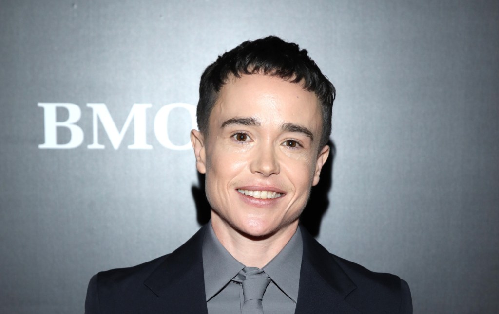 Elliot Page se tient sur un fond gris. Il porte une chemise grise et une cravate avec un blazer noir. Souriant devant la caméra.