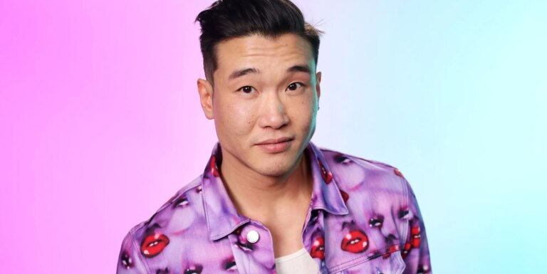 Joel Kim Booster affirme que les « relations sexuelles récréatives » en dehors de son mariage ouvert ajoutent de la pression Joel Kim Booster in a jacket with purple faces and red lips. The background is hazy pink and blue.