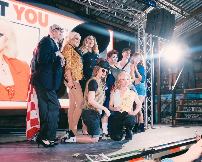 Un groupe d'artistes de drag post sur scène devant un grand écran de projection montrant une image promotionnelle de l'événement.