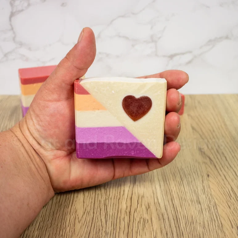 une main tenant un pain de savon artisanal conçu aux couleurs du drapeau de la fierté lesbienne
