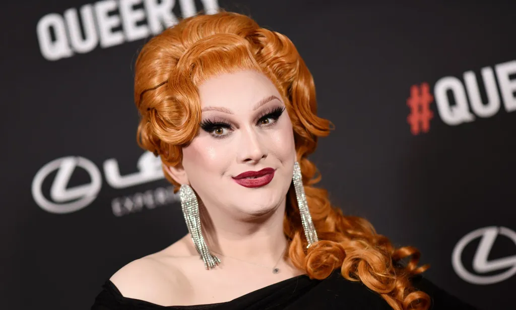 Jinkx Monsoon portant une robe noire et des boucles d'oreilles argentées sur un tapis rouge.