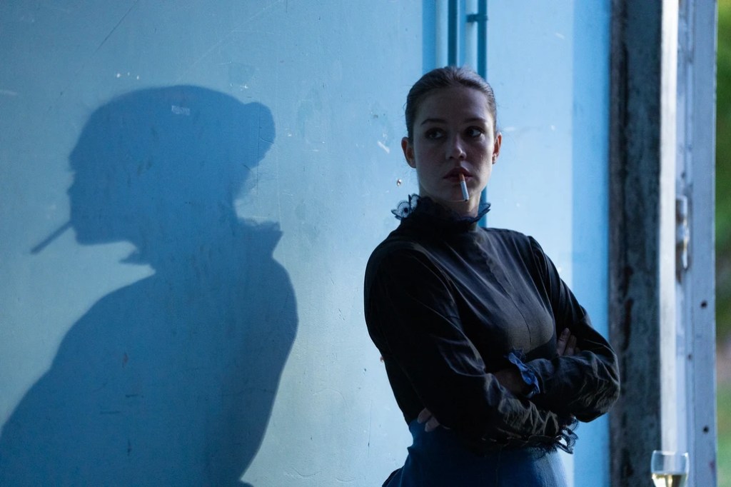 Another Day still : une femme qui fume, derrière elle sur un mur bleu son ombre étendue