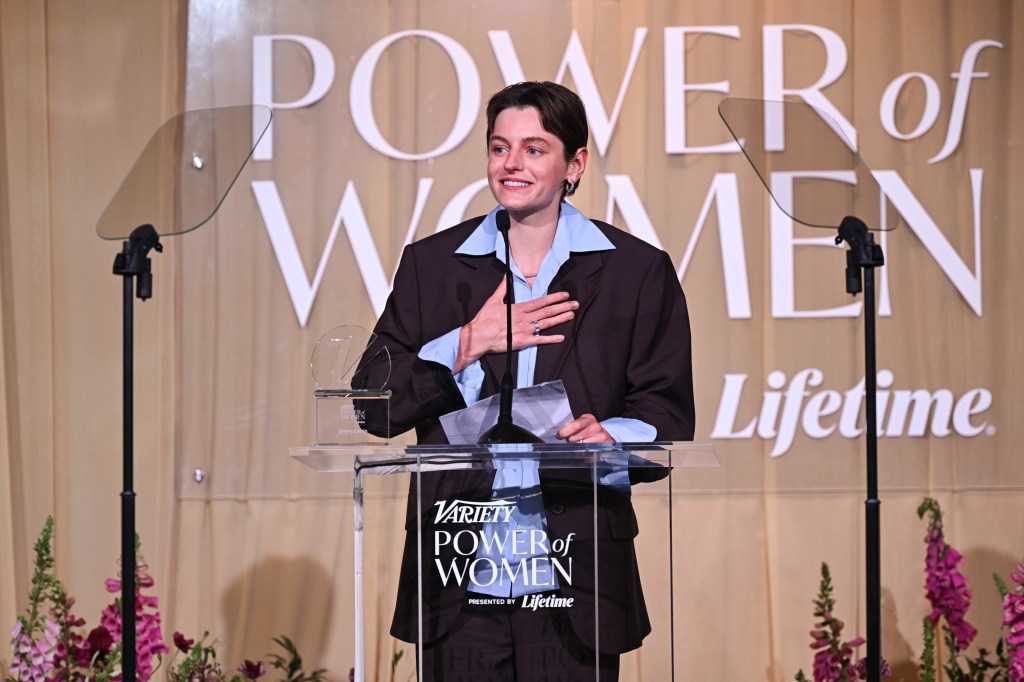 Emma Corrin à Variety 2026 Power of Women Londres. Ils portent une chemise bleu pâle sous un costume marron, ils sont debout sur un podium en train de lire leur discours.