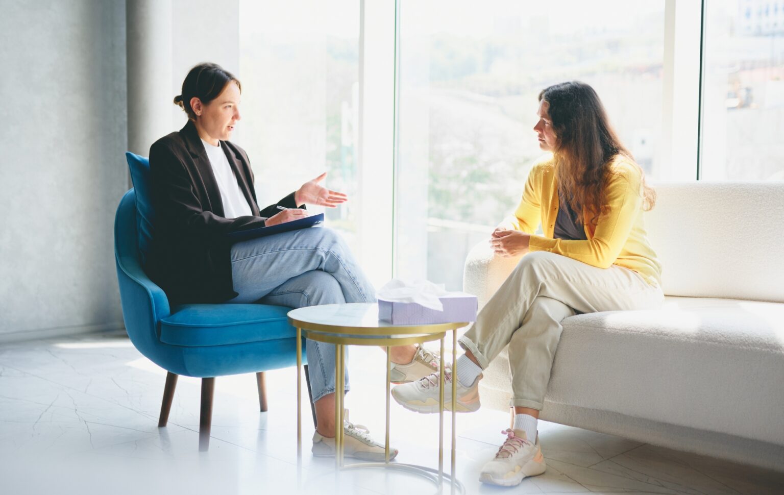 Un organisme britannique majeur reconnaît ses échecs en s'engageant en faveur des personnes trans Two women are seated in a bright room with large windows. One woman, wearing a yellow sweater, is listening attentively while the other woman takes notes. The setting is professional, suggesting a counseling or meeting scenario. The room features a blue chair, a white sofa, and a small table with a tissue box. No visible copy space.