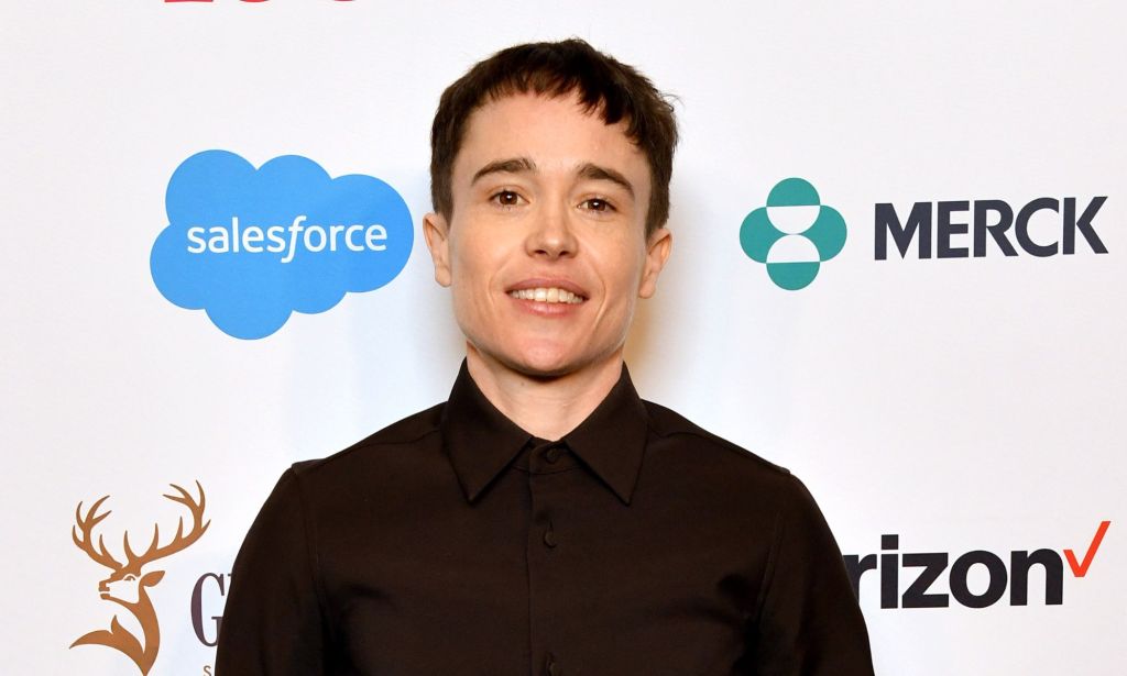 Elliot Page sourit sur un tapis rouge tout en portant une chemise boutonnée sombre.