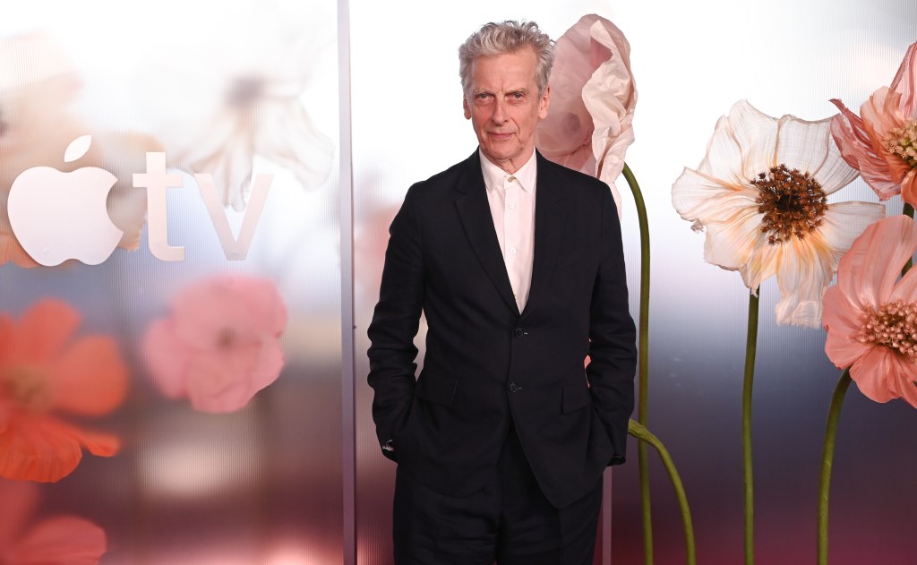 Peter Capaldi en costume noir devant un mur de coquelicots