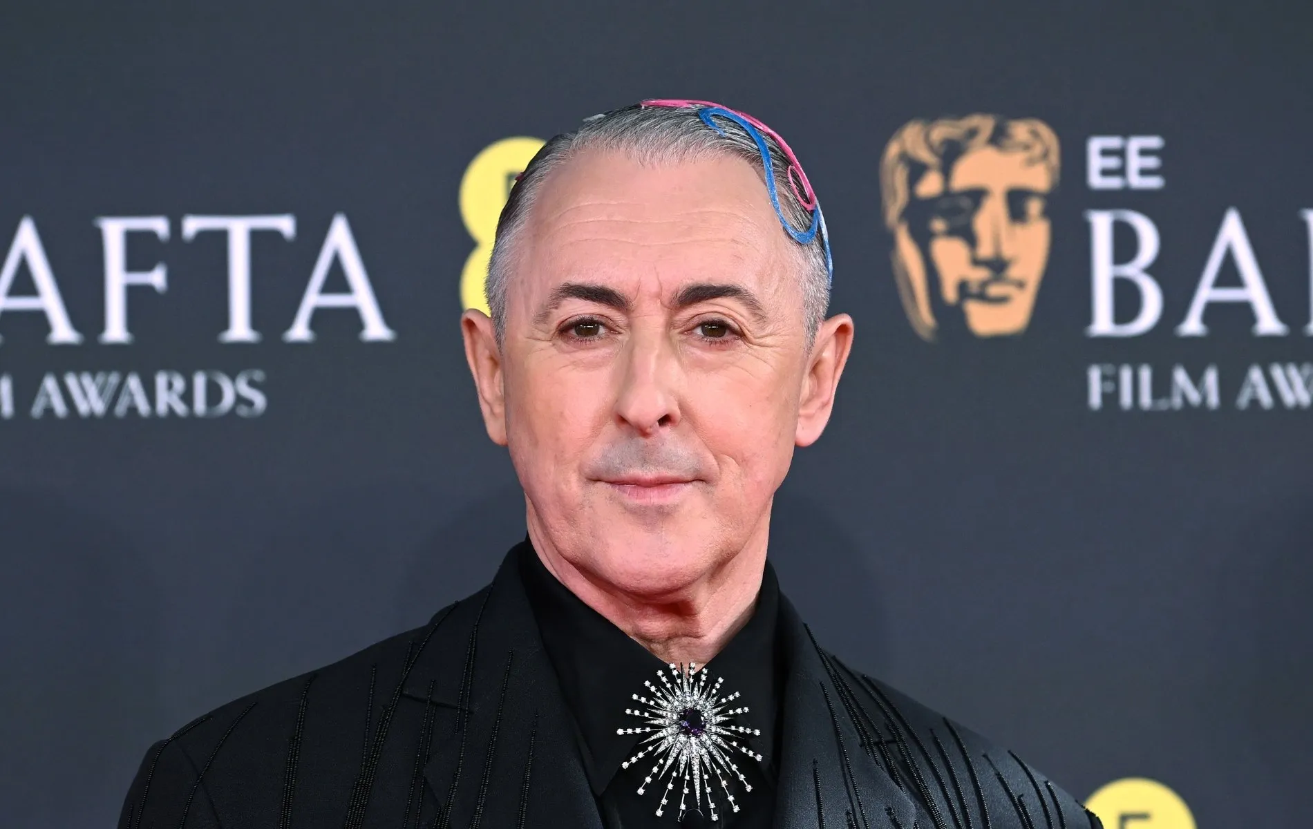 Alan Cumming assiste aux EE BAFTA film Awards 2026 au Royal Festival Hall. Il porte une veste de costume noire avec des accessoires pour cheveux roses et bleus. Une broche en argent sur son noeud de cravate